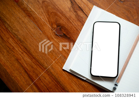 Top view of a workspace with a white-screen smartphone mockup on a book on a wooden desk. Top view of a workspace with a white-screen smartphone mockup on a book on a wooden desk. 108328941