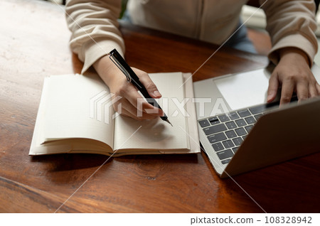 Cropped image a woman working on her laptop and taking notes in her notebook at a desk. Cropped image a woman working on her laptop and taking notes in her notebook at a desk. 108328942