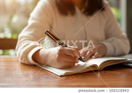 Cropped image of an Asian woman in casual clothes writing something in her book at a table indoors. Cropped image of an Asian woman in casual clothes writing something in her book at a table indoors. 108328943