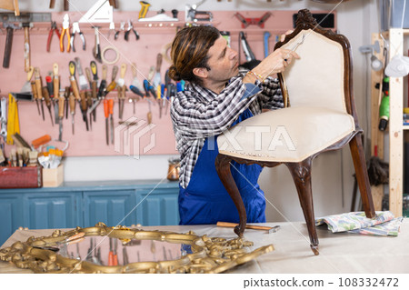 Middle-aged male restorer removes old cloth seat covering from antique wooden chair. 108332472