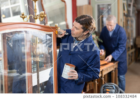 middle-aged furniture maker applies varnish with brush to wooden parts of vintage cabinet 108332484