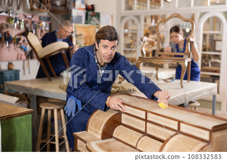 Professional middle-aged craftsman restorer in uniform renewing antique dresser while working in repair shop 108332583