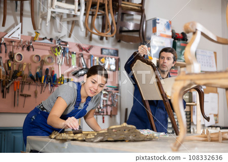 Girl restorer carefully covers carved wooden frame of antique wall mirror with thin layer of varnish 108332636