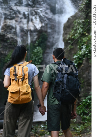 Rear view of loving couple tourist with backpack admiring a beautiful waterfall Rear view of loving couple tourist with backpack admiring a beautiful waterfall 108336155