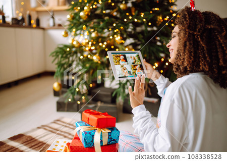 Young woman on background of Christmas tree with gifts, with tablet has video call or video chat. 108338528