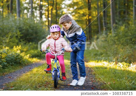 Cute little preschool girl in safety helmet riding bicycle. School kid boy, brother teaching happy healthy sister child cycling and having fun with learning bike. Active siblings family outdoors. 108338715