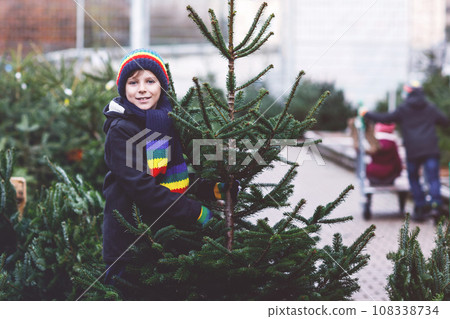 Adorable little smiling kid boy holding Christmas tree on market. Happy healthy child in winter fashion clothes choosing and buying big Xmas tree in outdoor shop. Family, tradition, celebration. 108338734