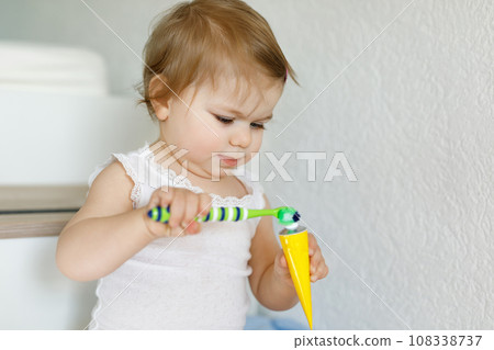 Little baby girl holding toothbrush and brushing first teeth. Toddler learning to clean milk tooth. Prevention, hygiene and healthcare concept. Happy child in bathroom 108338737
