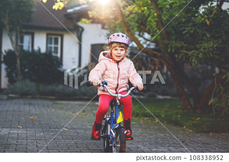 Little preschool girl riding bike. Kid on bicycle outdoors. Happy child enjoying bike ride on her way to school on warm summer day. Preschooler learning to balance on bicycle in safe helmet. 108338952