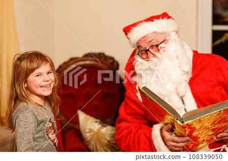 Little toddler girl talking to Santa Claus called Nikolaus or Weihnachtsmann in German. Happy smiling kid waiting for gifts. Santa with golden book. Xmas, childhood, holiday 108339050