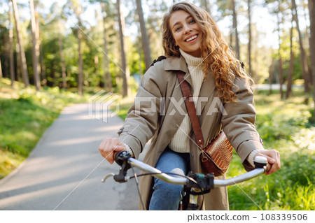 Beautiful tourist woman with curls in coat rides bicycle enjoying spring weather. Active lifestyle. Beautiful tourist woman with curls in coat rides bicycle enjoying spring weather. Active lifestyle. 108339506