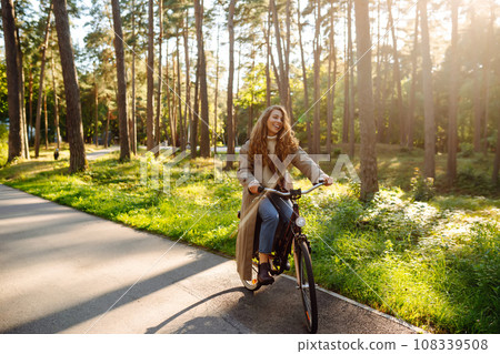 Beautiful tourist woman with curls in coat rides bicycle enjoying spring weather. Active lifestyle. 108339508