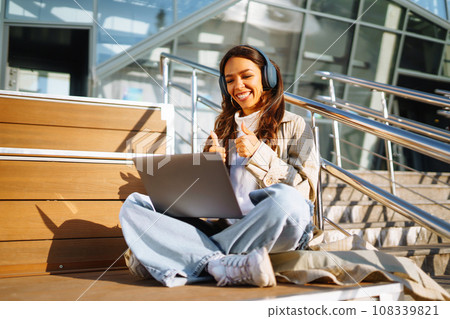 Young woman in headphones with laptop on city street. Video conference. ?nline training. Freelancer 108339821