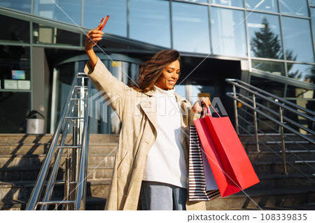 Young woman standing along road holding shopping bags, using mobile phone. Discounts, sale concept. 108339835