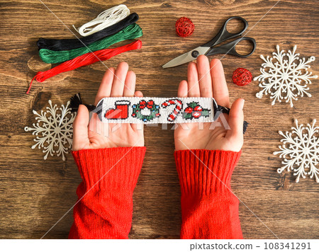 Idea for Christmas gift. Friendship bracelet with New Year alpha pattern handmade of floss threads in female hands in red sweater. Scissors, skeins of embroidery thread on wooden background.  108341291