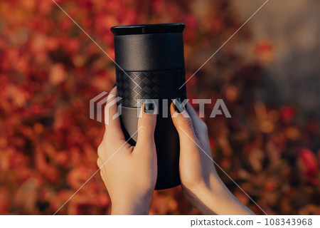 Woman hands holds black thermos cup with fresh coffee. Drinking in autumn forest 108343968