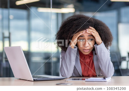 Frustrated and tired depressed business woman sitting at workplace inside office, unhappy african american woman in business suit working with laptop, failed and bankrupt. Frustrated and tired depressed business woman sitting at workplace inside office, unhappy african american woman in business suit working with laptop, failed and bankrupt. 108345200