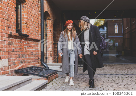 Two girls walking on street together and holding their hands. They are wearing spring or autumn clothes and they are happy. Friendship and relationship concept 108345361