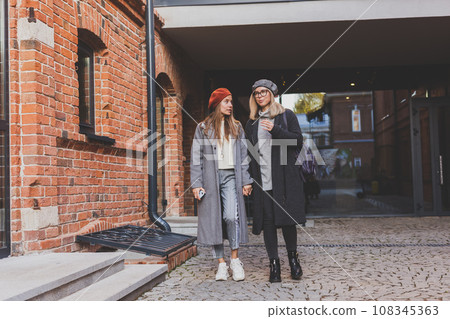 Two girls walking on street together and holding their hands. They are wearing spring or autumn clothes and they are happy. Friendship and relationship concept 108345363