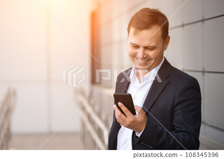 Adult smiling businessman in suit writes message on phone Adult smiling businessman in suit writes message on phone 108345396
