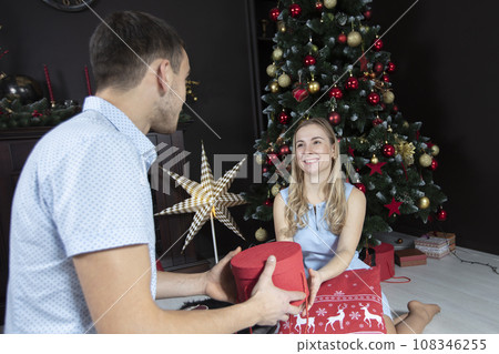 A man holds a red gift box on the background of his wife and child. Family on New Year's holidays. The little son runs to receive a Christmas gift. 108346255