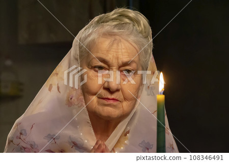 An elderly woman in a headscarf looks at a candle.An elderly woman is looking at a candle. The old woman is praying. 108346491