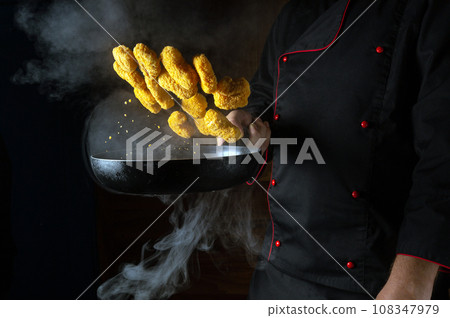 The hand of a professional chef throws nagits in a hot frying pan with steam on a black background. The concept of cooking in the hotel. Free ad space The hand of a professional chef throws nagits in a hot frying pan with steam on a black background. The concept of cooking in the hotel. Free ad space 108347979