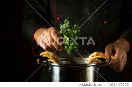 A cook in a black uniform adds parsley to a pan of boiling chicken legs. Preparing a delicious soup or fatty side dish in the kitchen. Cooking concept with chicken or rooster on black background A cook in a black uniform adds parsley to a pan of boiling chicken legs. Preparing a delicious soup or fatty side dish in the kitchen. Cooking concept with chicken or rooster on black background 108347995