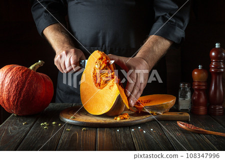 A chef cuts a pumpkin with a kitchen knife on a wooden board in a restaurant kitchen. The process of preparing ingredients for a seasonal fall dish, soup, or national American pie A chef cuts a pumpkin with a kitchen knife on a wooden board in a restaurant kitchen. The process of preparing ingredients for a seasonal fall dish, soup, or national American pie 108347996