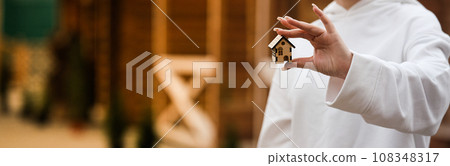 A small wooden house on a woman s hand against the background of a built wooden house. Concept of buying an ecological house made of wood 108348317