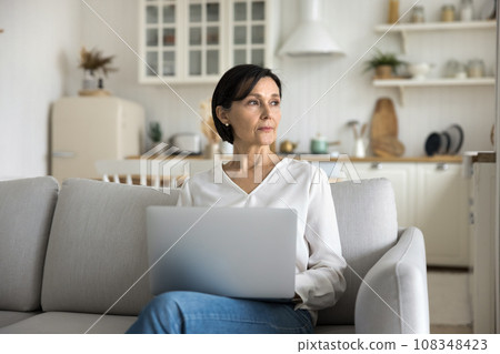 Serious mature female sit on sofa with computer on laps Serious mature female sit on sofa with computer on laps 108348423