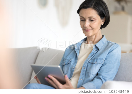 Mature woman working on tablet seated on cozy couch Mature woman working on tablet seated on cozy couch 108348449
