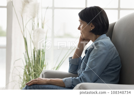 Pensive mature woman sits on armchair staring into distance Pensive mature woman sits on armchair staring into distance 108348734