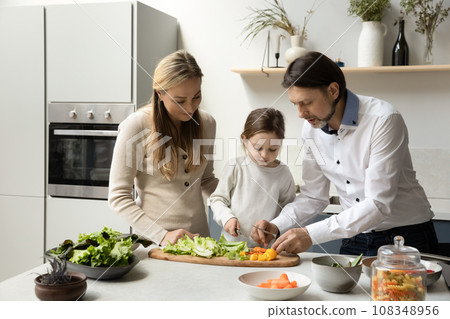 Focused daughter kid helping mom and dad to cook salad Focused daughter kid helping mom and dad to cook salad 108348956