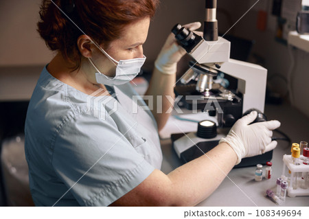 Middle aged lab technician adjusts microscope to research material sample at workplace 108349694