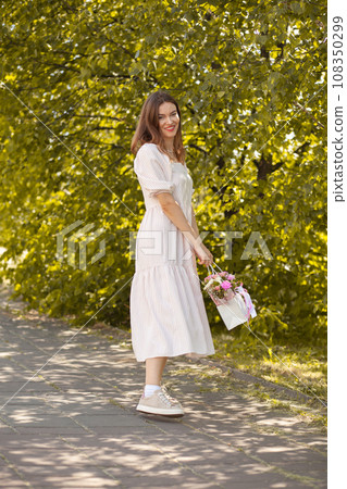 Beautiful girl with flowers, in a dress, glasses and sneakers. A beautiful bouquet of flowers in a box in the hands of a beautiful girl who walks along the street on a sunny day. Beautiful girl with flowers, in a dress, glasses and sneakers. A beautiful bouquet of flowers in a box in the hands of a beautiful girl who walks along the street on a sunny day. 108350299