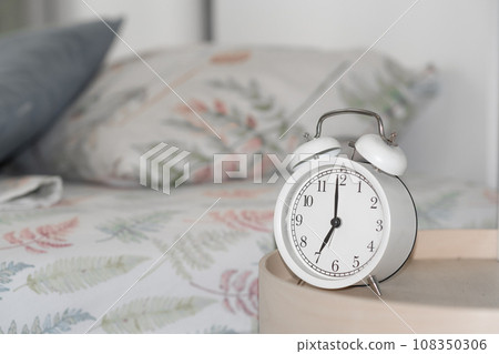 Close-up of a round white alarm clock on a table in the bedroom. The hands on the clock show seven o'clock in the morning, time to get up. Retro alarm clock on the table, vintage tone. space for text Close-up of a round white alarm clock on a table in the bedroom. The hands on the clock show seven o'clock in the morning, time to get up. Retro alarm clock on the table, vintage tone. space for text 108350306