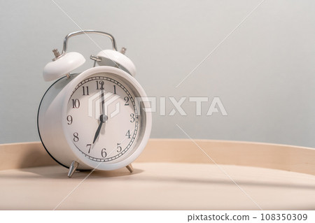 Close-up of a round white alarm clock on a table in the bedroom. The hands on the clock show seven o'clock in the morning, time to get up. Retro alarm clock on the table, vintage tone. space for text Close-up of a round white alarm clock on a table in the bedroom. The hands on the clock show seven o'clock in the morning, time to get up. Retro alarm clock on the table, vintage tone. space for text 108350309