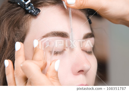 Painting eyebrows with an eyebrow brush. Eyebrow care, eyebrow lamination. close-up of the master's hands with a microbrush, which combs the model's eyebrows after the procedure 108351787