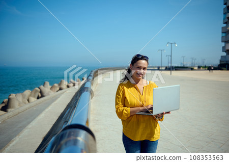Happy multi-ethnic business woman smiling broadly, looking at camera, standing with laptop along the coastline promenade Happy multi-ethnic business woman smiling broadly, looking at camera, standing with laptop along the coastline promenade 108353563