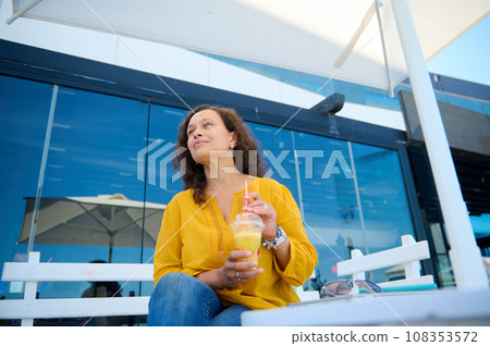 View from the bottom of a young adult pretty woman in casual clothes, sitting on a cafe, holding a glass of fresh juice 108353572