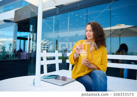 Multi-ethnic young adult woman holding glass of freshly squeezed orange juice, looking dreamily aside, sitting on a cafe Multi-ethnic young adult woman holding glass of freshly squeezed orange juice, looking dreamily aside, sitting on a cafe 108353573