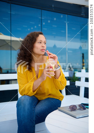Attractive brunette sitting in cafe with laptop on table, looks thoughtfully aside and takes a sip of fresh orange juice Attractive brunette sitting in cafe with laptop on table, looks thoughtfully aside and takes a sip of fresh orange juice 108353574