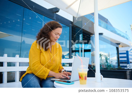Successful adult female freelancer typing on laptop, while working remotely at table with juice glass in outdoor cafe Successful adult female freelancer typing on laptop, while working remotely at table with juice glass in outdoor cafe 108353575