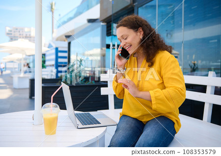 Beautiful ethnic middle aged woman with refreshing drink and laptop at outdoor cafe, smiling talking on mobile phone 108353579
