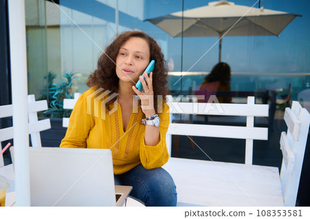 Multitasking businesswoman talking on mobile phone, working remotely on laptop from a modern outdoor cafe Multitasking businesswoman talking on mobile phone, working remotely on laptop from a modern outdoor cafe 108353581