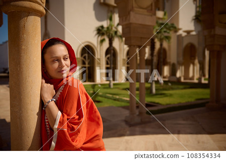 Attractive woman in Oriental authentic dress and head covered in hijab, leaning on a marble column in the mosque garden 108354334