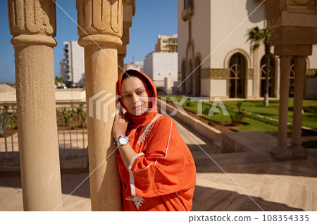 Stylish elegant Arab Muslim woman in Moroccan ethnic orange dress, leaning on a marble column and dreamily looking away 108354335