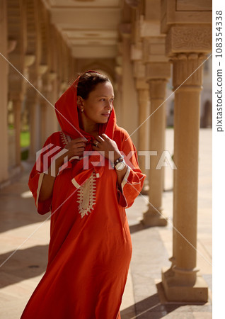 Beautiful young brunette woman in orange ethnic dress and head covered in hijab, posing against the mosque background Beautiful young brunette woman in orange ethnic dress and head covered in hijab, posing against the mosque background 108354338