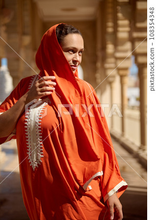 Dynamic fashion portrait of a beautiful young adult woman in Arabian attire and head covered in a shawl, looking aside 108354348